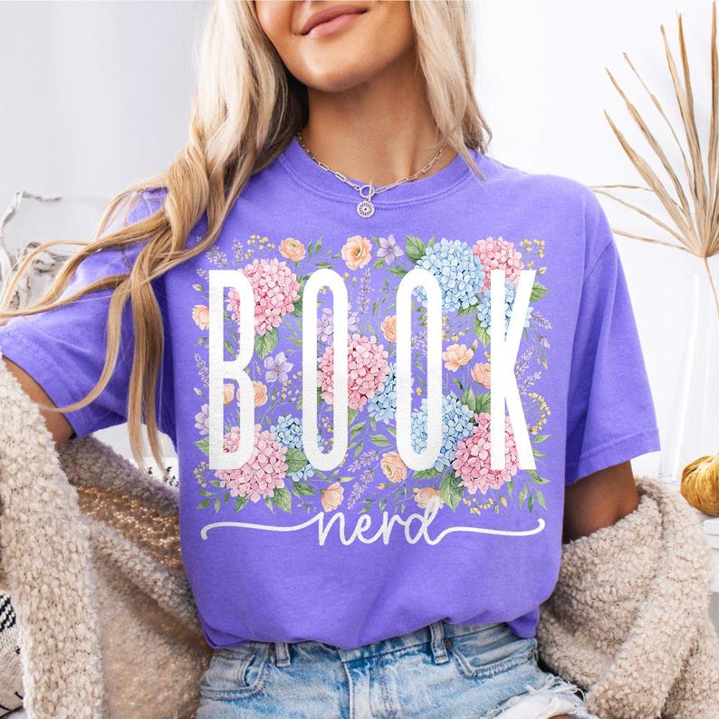 May include: A lilac t-shirt featuring the text "BOOK nerd" in white, set against a floral arrangement of pink, blue, and peach blooms. The shirt is a short-sleeved, casual style.