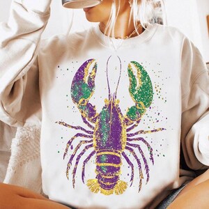 May include: A white sweatshirt with a purple, green, and gold glitter lobster design. The lobster is in a standing position with its claws raised.