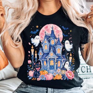 Puede incluir: Una camiseta negra con un estampado gr&aacute;fico de una casa encantada azul bajo una gran luna rosa. El dise&ntilde;o incluye fantasmas blancos, un gato negro con un lazo rosa, calabazas naranjas, velas encendidas, murci&eacute;lagos y flores coloridas.
