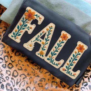 May include: A dark gray sweatshirt with the word "FALL" embroidered in cream-colored letters. Each letter is decorated with orange, yellow, and green floral designs. The sweatshirt is folded on a leopard print surface.