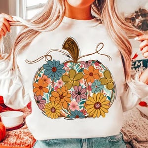 May include: White long-sleeved sweatshirt with a floral pumpkin design. The pumpkin is filled with colourful flowers in shades of orange, yellow, blue, and pink. The design has a brown stem and vines.