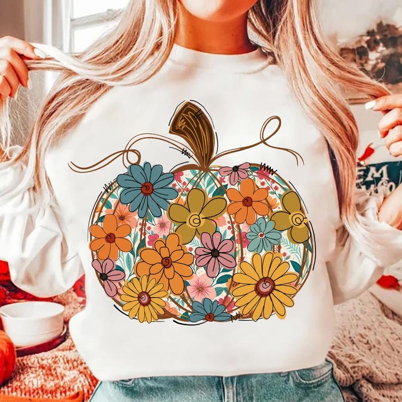 May include: White long-sleeved sweatshirt with a floral pumpkin design. The pumpkin is filled with colourful flowers in shades of orange, yellow, blue, and pink. The design has a brown stem and vines.