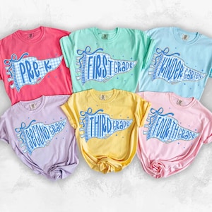 May include: Six short-sleeved t-shirts in various pastel colours, each featuring a blue and white gingham pennant graphic. The shirts display the text Pre-K, First Grade, Kindergarten, Second Grade, Third Grade, and Fourth Grade in blue script.