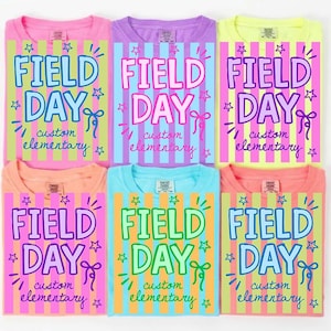 May include: Six colorful t-shirts with the words "FIELD DAY" in large, playful lettering. Each shirt has a different background color and stripe pattern, with the text "custom elementary" below. The shirts are in shades of pink, purple, yellow, orange, and blue.