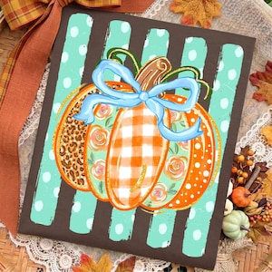May include: A rectangular sign featuring a brown background with vertical light blue polka-dot stripes. Centered is a pumpkin illustration with segments of leopard print, orange gingham, and floral patterns, tied with a light blue ribbon bow.