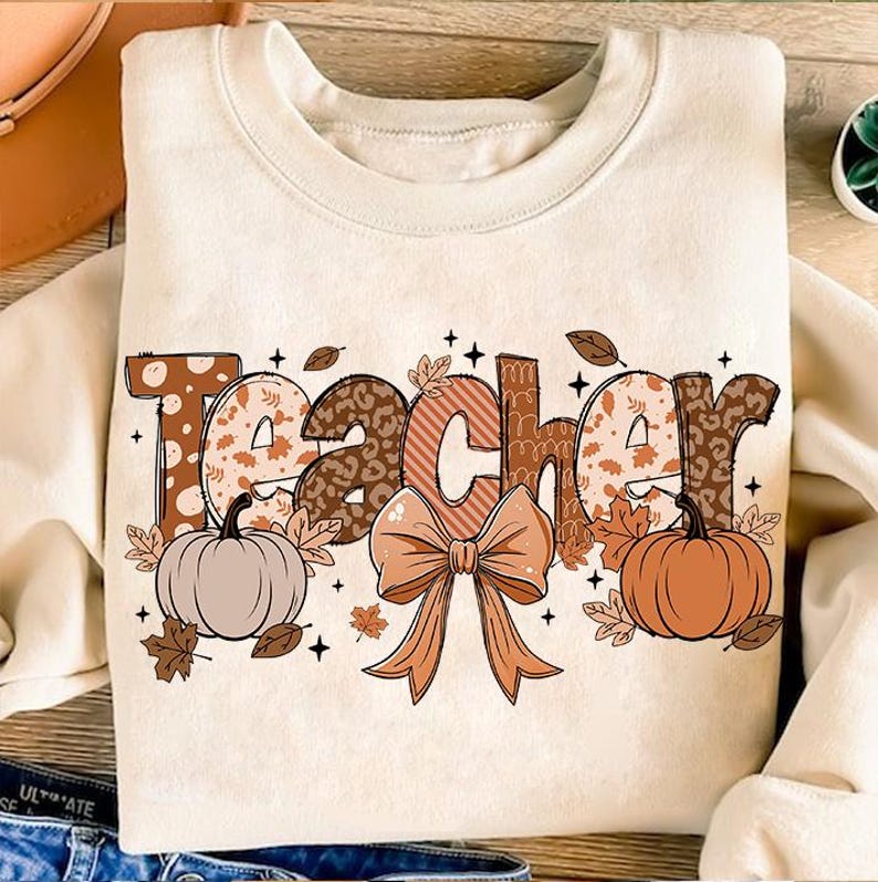 May include: A beige sweatshirt with the word "Teacher" printed in brown, orange, and white letters. The letters are decorated with fall-themed patterns, including leaves, pumpkins, and a bow.