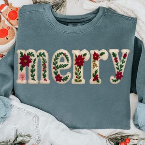 May include: A teal sweatshirt featuring the word "MERRY" in cream-coloured, embroidered letters. Each letter is adorned with red poinsettias, green leaves, and pink flowers, creating a festive, seasonal design.
