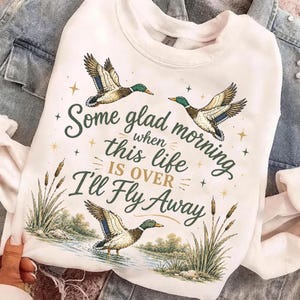 May include: White sweatshirt with a graphic of three ducks in flight and one duck in water. The text reads "Some glad morning when this life is over I'll Fly Away" in a script font. The design includes cattails and stars.