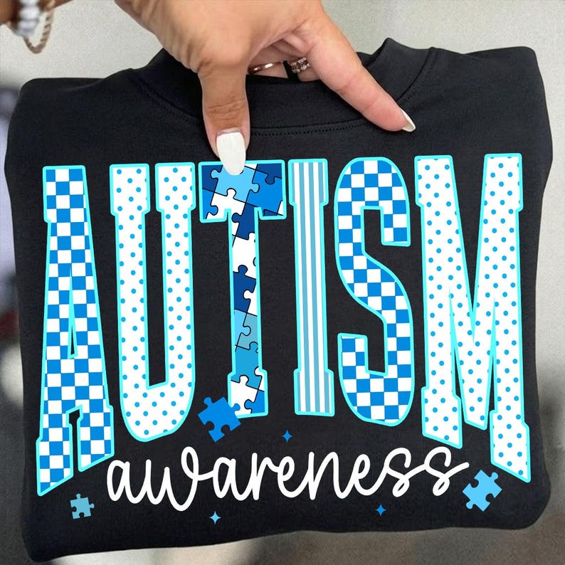 May include: Black sweatshirt with the words "AUTISM awareness" in white and blue. The word "AUTISM" is in a mix of checkered, dotted, and puzzle piece designs. The word "awareness" is in a cursive font.