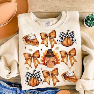 Puede incluir: Sudadera de color crema con un diseño temático de Acción de Gracias. El diseño incluye ilustraciones de rebanadas de pastel de calabaza, lazos naranjas, calabazas con patrones de cuadros y un pavo de dibujos animados.