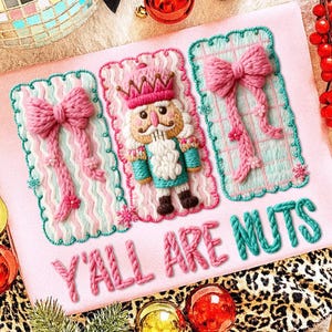 May include: Embroidered pink fabric with a festive design. It features a nutcracker, bows, and the text "Y'ALL ARE NUTS". The design incorporates pink, teal, and white, with a holiday theme, perfect for Christmas.