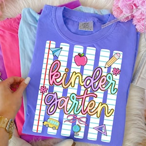 Peut inclure: Un t-shirt lilas Comfort Colors avec un motif "Kindergarten" color&eacute;. Le motif comprend un avion en papier, une pomme, un crayon, une fleur, un bus scolaire, un n&oelig;ud, un globe et un triangle. Le t-shirt est pli&eacute; avec d'autres t-shirts aux couleurs pastel.