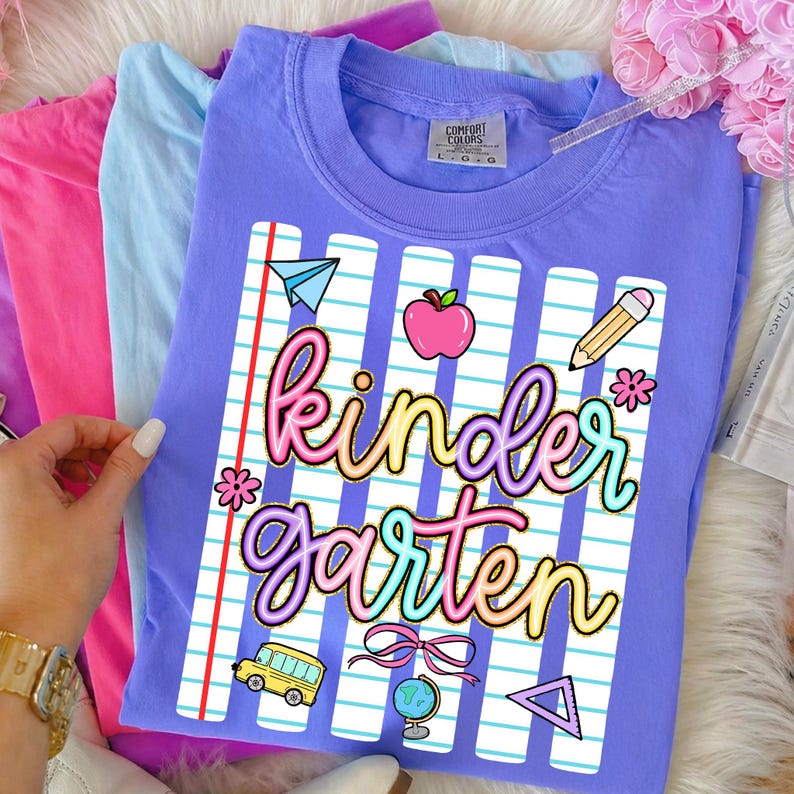 Peut inclure: Un t-shirt lilas Comfort Colors avec un motif "Kindergarten" color&eacute;. Le motif comprend un avion en papier, une pomme, un crayon, une fleur, un bus scolaire, un n&oelig;ud, un globe et un triangle. Le t-shirt est pli&eacute; avec d'autres t-shirts aux couleurs pastel.