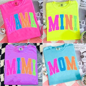 Może przedstawiać: Cztery kolorowe koszulki z napisami "MAMA", "MINI", "MIMI" i "MOM" w stylu retro. Koszulki są w kolorze r&oacute;żowym, neonowo-ż&oacute;łtym, fioletowym i jasnoniebieskim. Tekst jest w kolorach tęczy.
