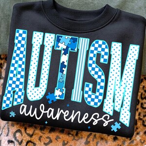 May include: Black sweatshirt with the word "AUTISM" in large, blue, patterned letters. The word "awareness" is written in white script below. The design includes puzzle pieces and stars, promoting autism awareness.