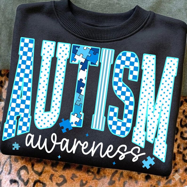 May include: Black sweatshirt with the word "AUTISM" in large, blue, patterned letters. The word "awareness" is written in white script below. The design includes puzzle pieces and stars, promoting autism awareness.