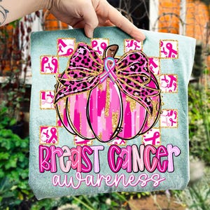 May include: A teal-colored tote bag featuring a pink pumpkin design with a leopard print bow and gold accents. The words "Breast Cancer Awareness" are written in pink and white. Pink ribbons are in the background.