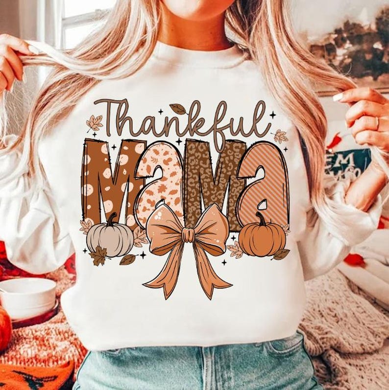 Op de afbeelding: Wit sweatshirt met een bruine en oranje grafische print die "Thankful Mama" zegt. De afbeelding bevat een strik, pompoenen en bladeren.