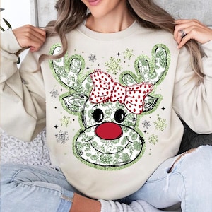 May include: A beige sweatshirt with a green and red reindeer design. The reindeer has a red nose, a red bow with white polka dots, and green floral details. The sweatshirt is decorated with snowflakes.
