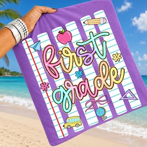Peut inclure: Une serviette de plage violette avec un motif sur le th&egrave;me de l'&eacute;cole. La serviette pr&eacute;sente les mots "First Grade" dans des lettres color&eacute;es et fantaisistes, ainsi que des illustrations de fournitures scolaires telles qu'un crayon, une pomme et un bus scolaire. Le fond a un motif de papier lign&eacute;.