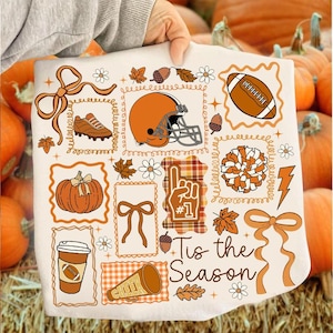 May include: A white decorative throw blanket featuring orange and brown fall-themed illustrations. The design includes a football, helmet, pumpkin, and the text "Tis the Season". The blanket is surrounded by pumpkins.