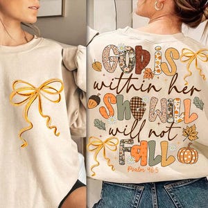 May include: Beige sweatshirt with a large gold bow graphic on the front. The back of the sweatshirt features the text "God is within her, she will not fall" with fall-themed illustrations, including leaves and a pumpkin.