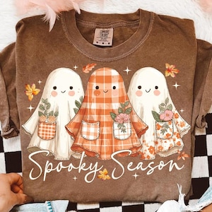 Könnte beinhalten: Braunes T-Shirt mit einer Grafik von drei lächelnden Geistern, jeder mit einer Tasche voller Blumen. Der mittlere Geist trägt ein orangefarbenes Outfit mit Karomuster. Der Text "Spooky Season" steht in weißer Schrift unten.