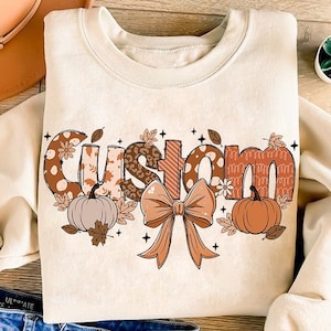 May include: Cream-colored sweatshirt with the word "Custom" in a fall-themed design. The letters feature patterns like polka dots, leopard print, and stripes, with pumpkins, leaves, and a bow. A seasonal autumn-themed apparel item.
