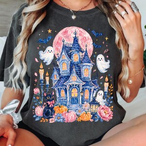 Puede incluir: Una camiseta gris carb&oacute;n con un estampado gr&aacute;fico de una casa encantada azul sobre una gran luna rosa. El dise&ntilde;o incluye fantasmas blancos, un gato negro con un lazo rosa, calabazas naranjas, velas encendidas y flores azules y rosas.