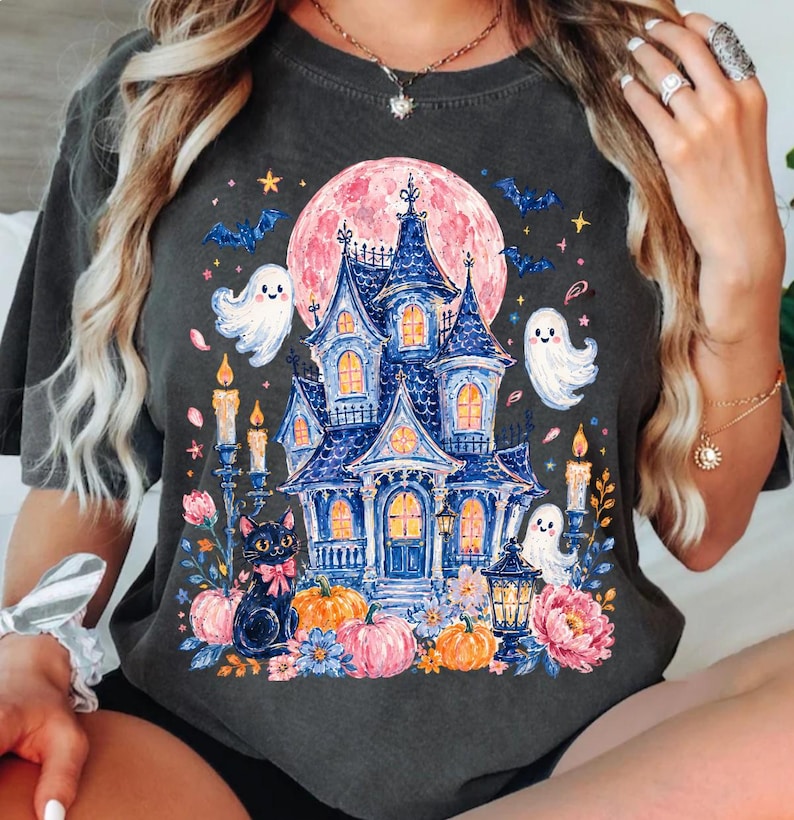 Puede incluir: Una camiseta gris carb&oacute;n con un estampado gr&aacute;fico de una casa encantada azul sobre una gran luna rosa. El dise&ntilde;o incluye fantasmas blancos, un gato negro con un lazo rosa, calabazas naranjas, velas encendidas y flores azules y rosas.