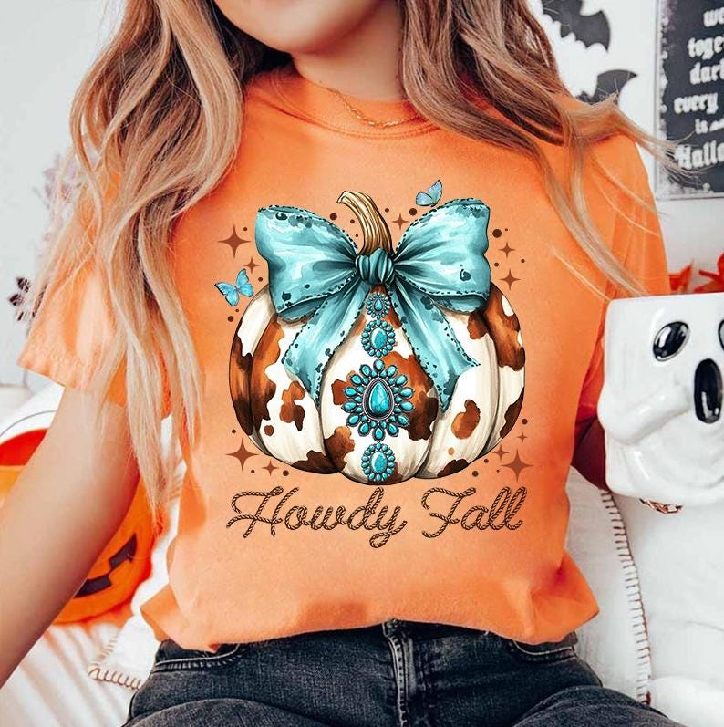 May include: Orange t-shirt with a brown and white cow print pumpkin with a turquoise bow and rhinestones. The text "Howdy Fall" is written in brown on a rope design.