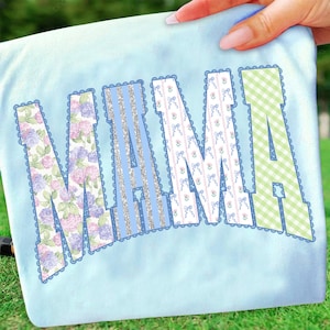 May include: Pale blue fabric featuring the word "NANA" in a decorative font. The letters are filled with floral, glitter, bow, and gingham patterns, with a scalloped border.