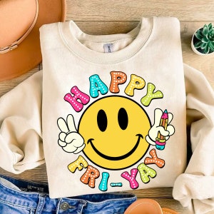 May include: A beige sweatshirt with a colorful graphic of a smiley face and the text "HAPPY FRI-YAY". The graphic features a peace sign, a pencil, and polka dots.