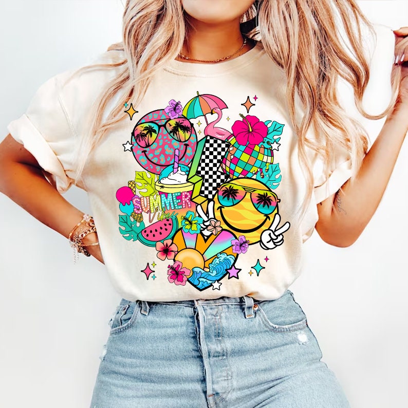 Peut inclure: Un t-shirt blanc avec un imprim&eacute; graphique color&eacute; sur le th&egrave;me de l'&eacute;t&eacute;. Le motif repr&eacute;sente des visages souriants avec des lunettes de soleil, des palmiers, une past&egrave;que, un flamant rose, un parapluie et le texte "Summer Vibes".