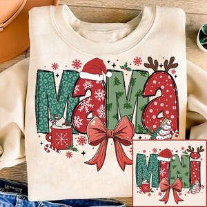 May include: A cream-colored sweatshirt with a Christmas-themed design. The words "Mama Mini" are written in a festive font with red, green, and white accents. The design includes a Santa hat, snowflakes, and a bow.