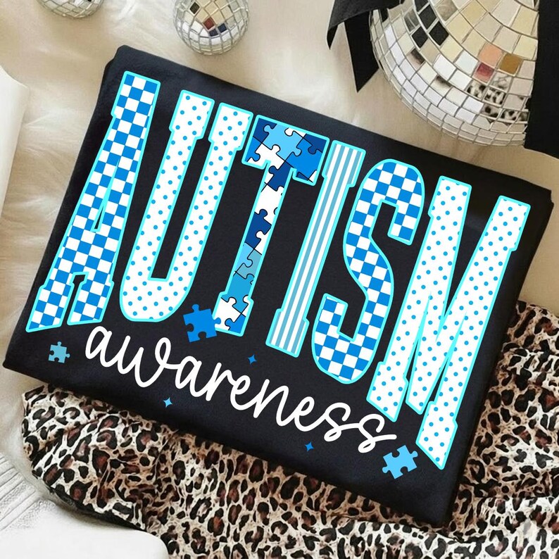 May include: Black t-shirt with the words "AUTISM awareness" in a blue and white design. The word "AUTISM" features puzzle piece details and checkered patterns. The shirt is folded on a leopard print fabric.