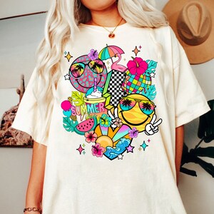 Peut inclure: Un t-shirt blanc avec un motif graphique color&eacute; repr&eacute;sentant deux visages souriants portant des lunettes de soleil, des palmiers, une past&egrave;que, un ananas, un flamant rose, un parasol de plage, un c&oelig;ur et le texte "SUMMER VIBES".
