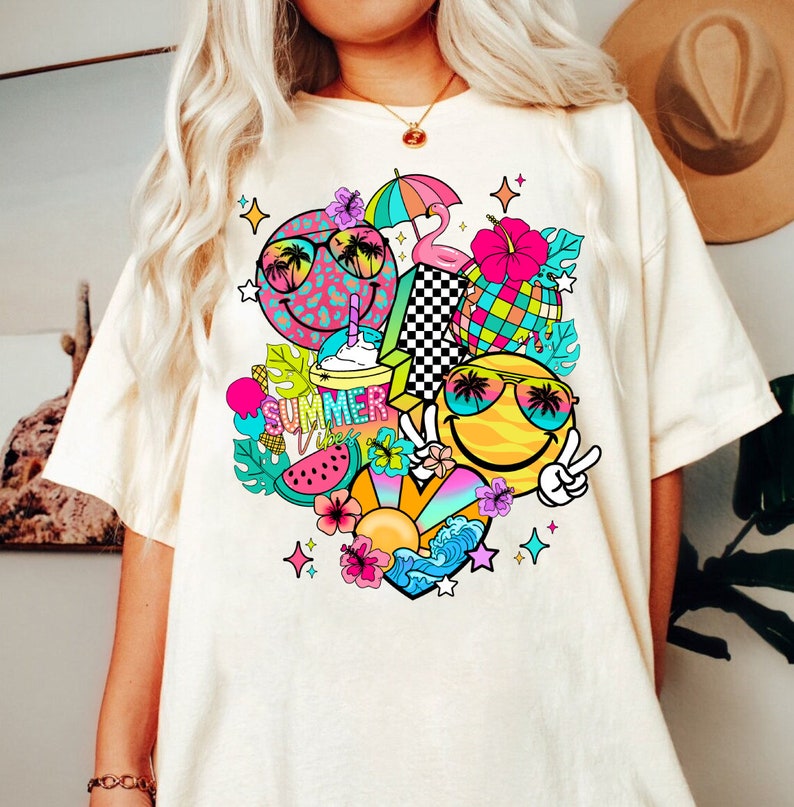 Peut inclure: Un t-shirt blanc avec un motif graphique color&eacute; repr&eacute;sentant deux visages souriants portant des lunettes de soleil, des palmiers, une past&egrave;que, un ananas, un flamant rose, un parasol de plage, un c&oelig;ur et le texte "SUMMER VIBES".