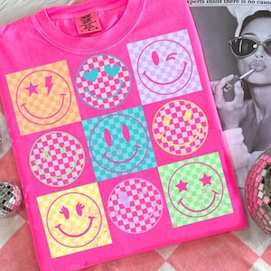 Könnte beinhalten: Leuchtend pinkes T-Shirt mit einem Raster aus Smileys auf kariertem Hintergrund in verschiedenen Farben. Jeder Smiley hat ein einzigartiges Design, einschließlich Herzen, Sternen und Blitzen. Das Shirt hat einen Rundhalsausschnitt und kurze Ärmel.