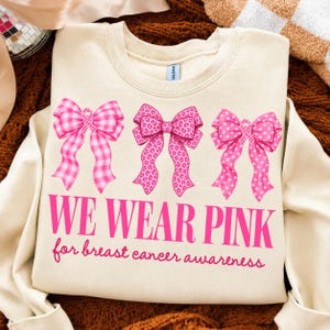 Op de afbeelding: Een crèmekleurige sweater met ronde hals voorzien van drie roze strikken met verschillende patronen: gingham, luipaardprint en stippen. Onder de strikken staat de tekst WE WEAR PINK for breast cancer awareness in opvallende roze letters.