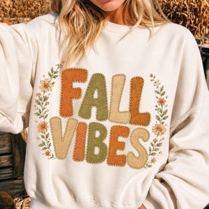 May include: A cream-colored crewneck sweatshirt featuring the text FALL VIBES in large, blocky, multi-colored letters with a stitched-look border. The text is surrounded by a decorative wreath of small orange and yellow flowers and green leaves.