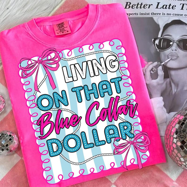 May include: A bright pink t-shirt with the phrase "LIVING ON THAT Blue Collar DOLLAR" in a decorative design. The design includes blue and white stripes, bows, and a pearl-like border. The shirt is made of a soft material.