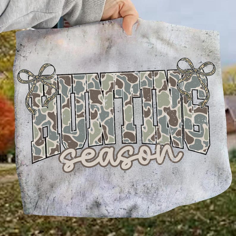 May include: A gray tote bag with the words "HUNTING season" in a camouflage pattern. The word "HUNTING" is in large block letters, and "season" is in a cursive font. The bag has two decorative bow accents.