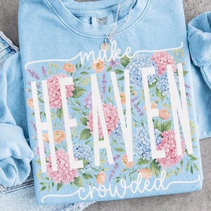 May include: Light blue sweatshirt with the words "make HEAVEN crowded" in white. The sweatshirt is decorated with a floral design of pink, blue, and peach flowers. A silver necklace with a small pendant is visible.
