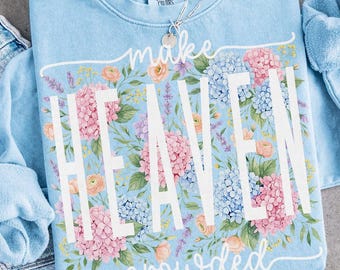 May include: Light blue sweatshirt with the words "make HEAVEN crowded" in white. The sweatshirt is decorated with a floral design of pink, blue, and peach flowers. A silver necklace with a small pendant is visible.