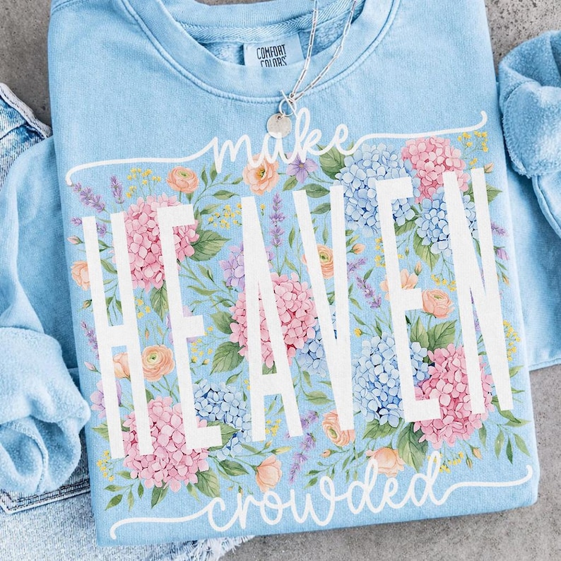 May include: Light blue sweatshirt with the words "make HEAVEN crowded" in white. The sweatshirt is decorated with a floral design of pink, blue, and peach flowers. A silver necklace with a small pendant is visible.