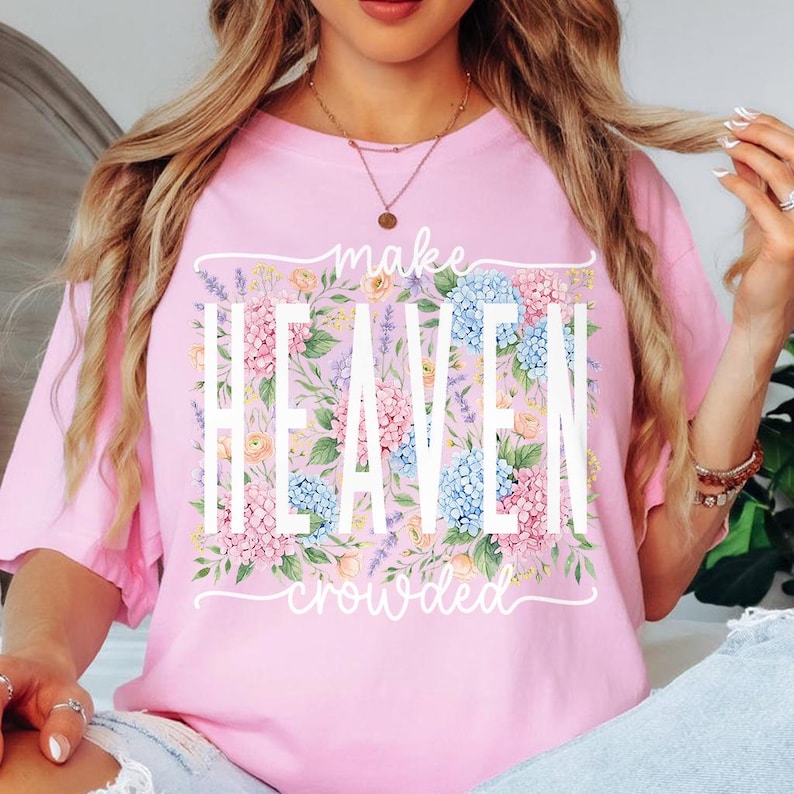 May include: Pink t-shirt with the words "make HEAVEN crowded" in white, surrounded by a floral design of pink, blue, and yellow flowers. The shirt is a solid pink color.