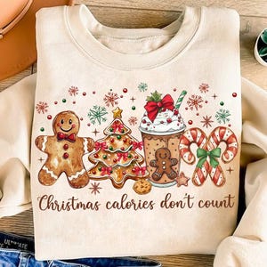 May include: Cream-colored sweatshirt with a festive Christmas graphic. The design features a gingerbread man, Christmas tree, coffee drink, and candy canes. The text "Christmas calories don't count" is printed below the graphic. Red, green, and white accents complete the holiday theme.