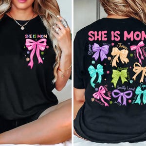 K&ouml;nnte beinhalten: Schwarzes T-Shirt mit dem Text "She is Mom" und einer rosa Schleife. Die R&uuml;ckseite des Shirts zeigt mehrere Schleifen in verschiedenen Farben mit Bibelversen und W&ouml;rtern wie "Chosen", "Victorious", "Capable" und "Greatly Loved".