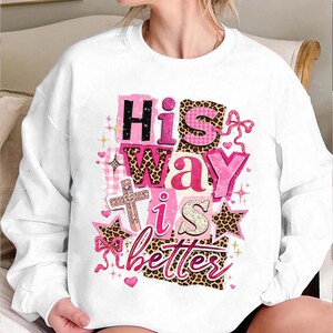 May include: White sweatshirt with the text "His Way is Better" in pink, black, and leopard print. The design includes a cross, stars, and bows. The sweatshirt is a casual, comfortable style.